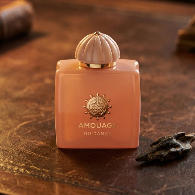 عطر زنانه آمواژ گایدنس ادوپرفیوم با شیشه صورتی کرم و درب گنبدی روی میز چوبی کلاسیک در کنار تکه چوب عود