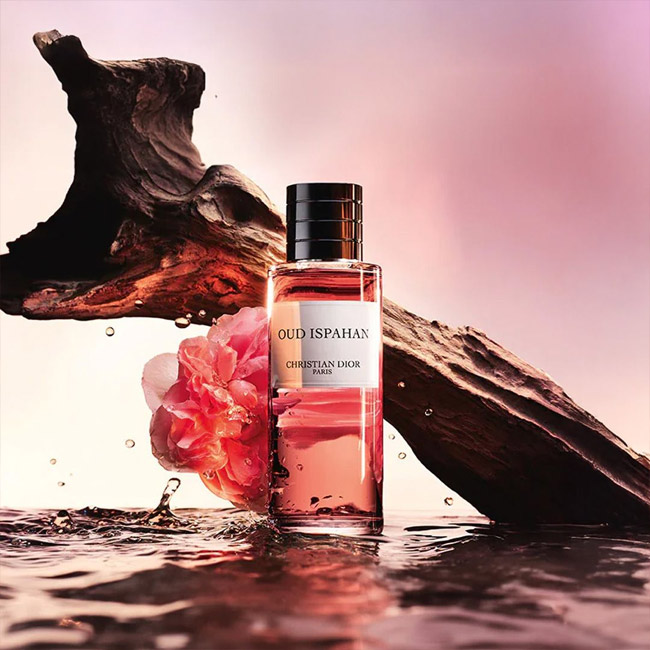 عطر دیور عود اسپهان اصل با شیشه شفاف و مایع صورتی در کنار تنه چوب عود و گل رز دمشقی روی سطح آب