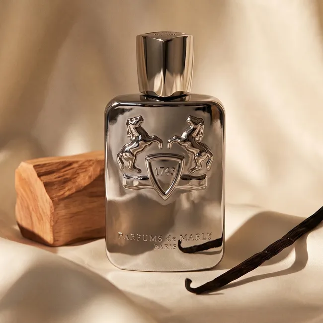 شیشه عطر پارفومز د مارلی پگاسوس نقره ای روی پارچه کرم کنار تکه چوب و وانیل