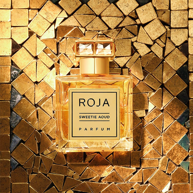 شیشه عطر روژا سویت عود با درپوش کریستالی روی پس زمینه کاشی های طلایی براق