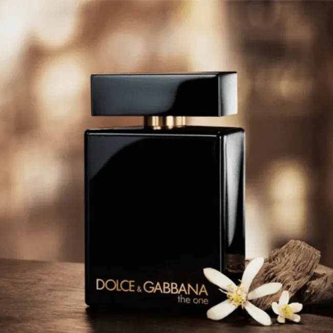 بطری عطر مشکی براق دولچه اند گابانا د وان روی میز چوبی، با دو گل سفید و چوب صندل در کنار آن